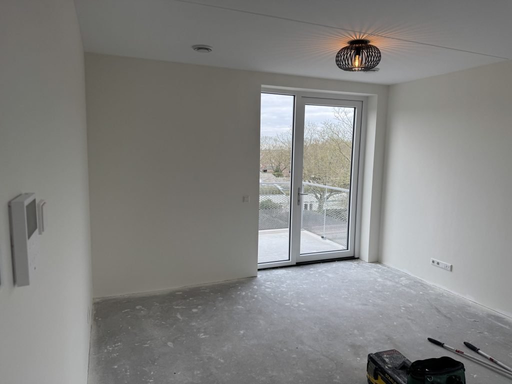 Lege kamer van een pas opgeleverd appartement met lichte wanden, een kale betonvloer en openslaande deuren naar het balkon